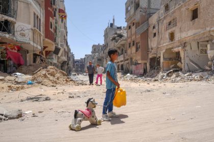 A Palestinian child stands in a rubble-strewn street in Gaza holding a yellow water jug in one hand and pulling a toy horse with the other.