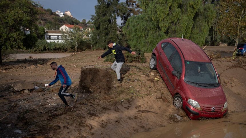 Men run next to a car covered with mud on a flooded street in Alora, near Malaga, on October 29, 2024, after a heavy rain hit southern Spain.