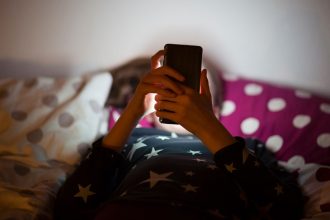 A girl lies in bed at night, scrolling through her phone.