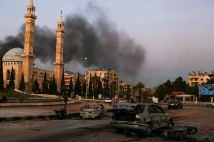 Smoke billows in the distance as damaged cars are seen at the site of Syrian regime airstrikes targeting opposition fighters in Aleppo, in northern Syria, on November 30, 2024.