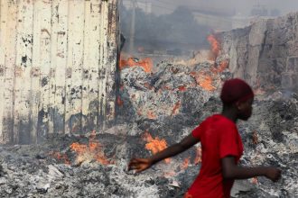 TOPSHOT - A woman walks past a fire at the burned down secondhand clothing market at Kantamanto in Accra, Ghana, on January 2, 2025. The fire at the Kantamanto market began in the early morning hours, destroying a large part of the area and displacing thousands of traders. (Photo by Nipah Dennis / AFP) (Photo by NIPAH DENNIS/AFP via Getty Images)