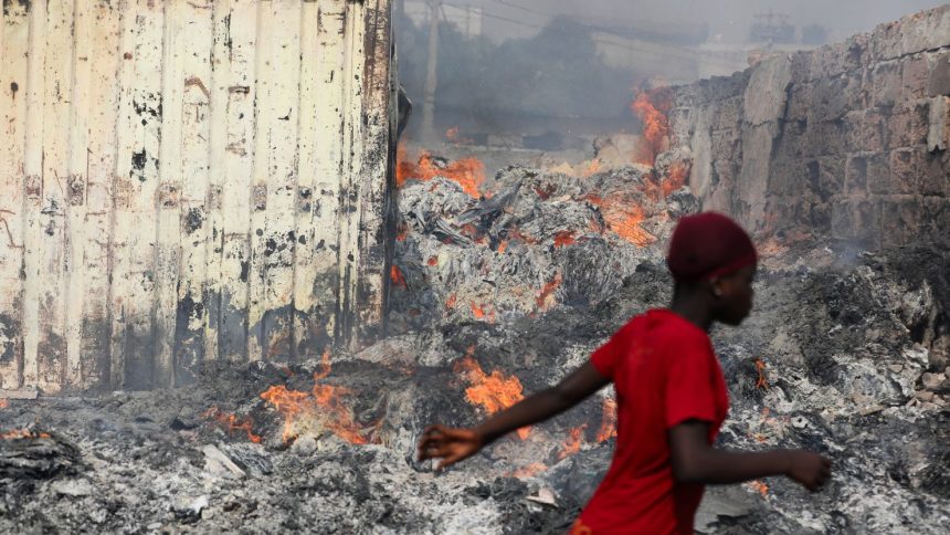 TOPSHOT - A woman walks past a fire at the burned down secondhand clothing market at Kantamanto in Accra, Ghana, on January 2, 2025. The fire at the Kantamanto market began in the early morning hours, destroying a large part of the area and displacing thousands of traders. (Photo by Nipah Dennis / AFP) (Photo by NIPAH DENNIS/AFP via Getty Images)