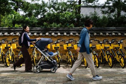 A woman pushes her child in a stroller past rental bicycles in Seoul on April 26, 2024.