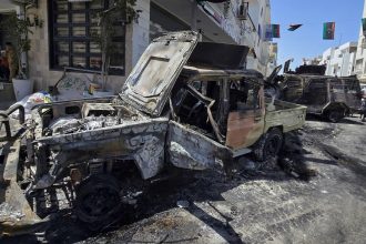 People sit at a coffee shop next to cars destroyed during overnight clashes in the southern district of Abu Salim in the Libyan capital Tripoli on May 13, 2025.